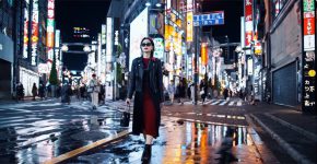 לכידת מסך מסרטון שנוצר לפי ההנחיות: אישה מסוגננת הולכת ברחוב בטוקיו מואר בניאון זוהר חמים ושילוט מונפש של העיר. היא לובשת ז'קט עור שחור, שמלה אדומה ארוכה ומגפיים שחורים.